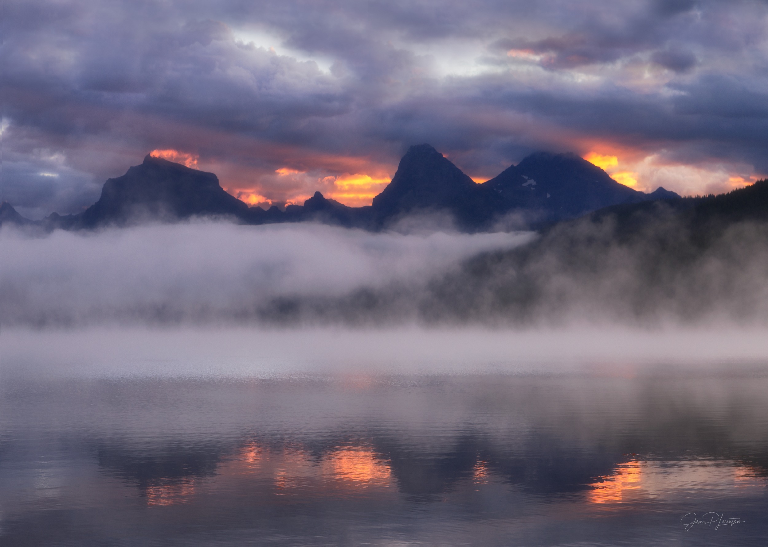 Steam fog drifting across a mountain lake at dawn while sunrise ignites the clouds behind dark peaks